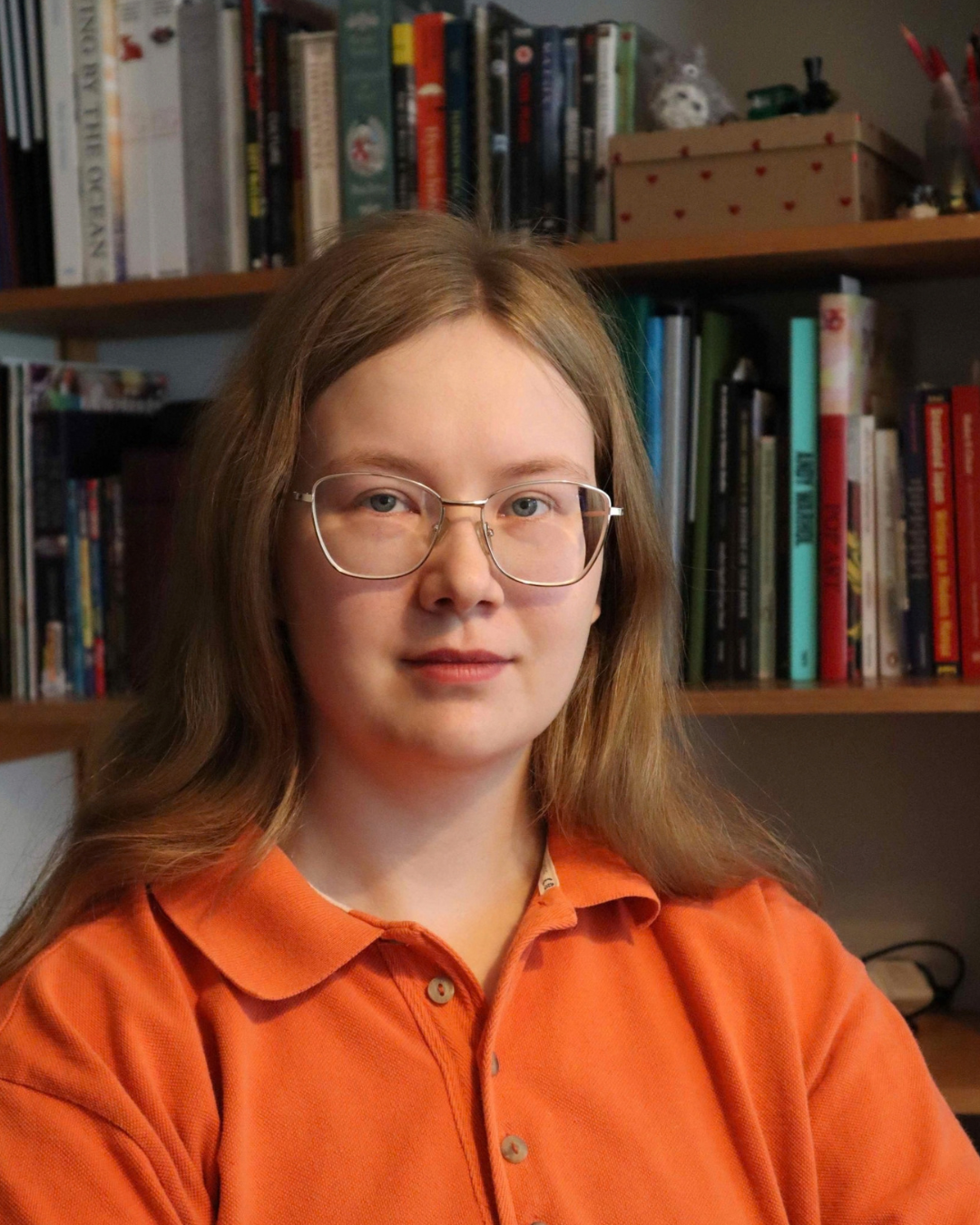 Portrait photo of Hanna, sitting in front of a bookshelf. She wears glasses and has orange shirt on. She looks content.